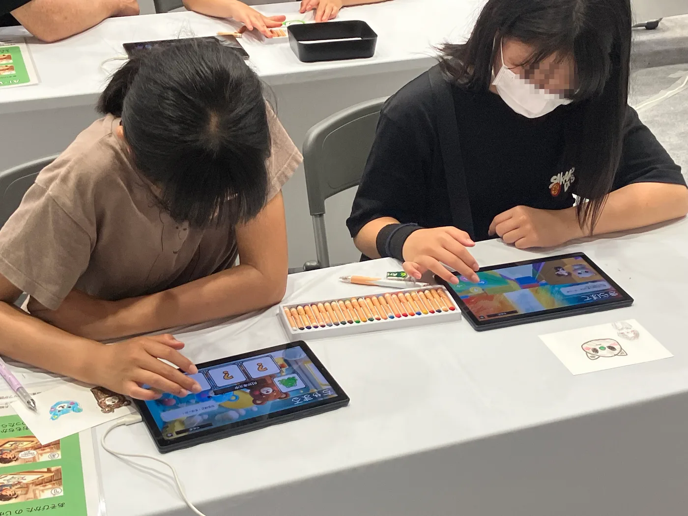 2人の子どもが隣り合ってタブレットで遊んでいる。テーブルの中央には色鉛筆のケース、手前には描いた猫の絵が置かれている