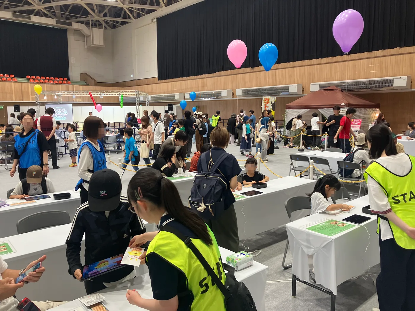 幌延町総合体育館の会場全景。風船が浮かび、多くの来場者が行き交うなか、白いテーブルが並ぶAI-KATAブースで子どもたちがタブレットに向かっている